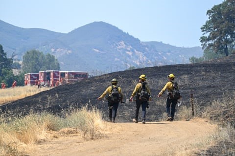 Three people in heavy yellow gear walk across burnt hillside toward red firetrucks, with hills beyond them.