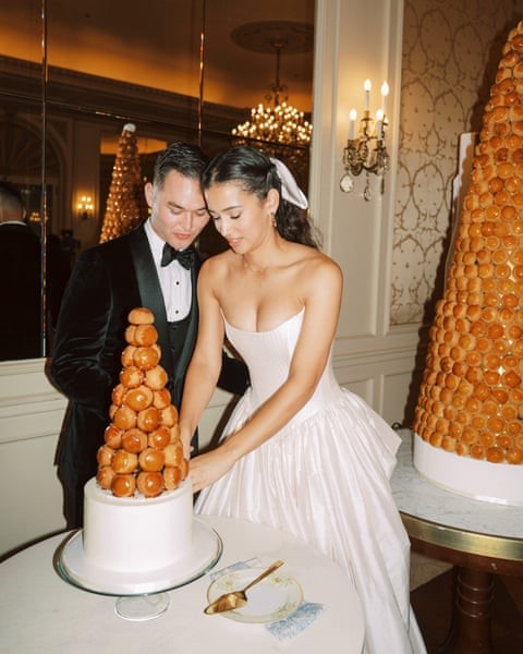 a bride and groom cutting a croquembouche cake. her dress is strapless with a drop waist