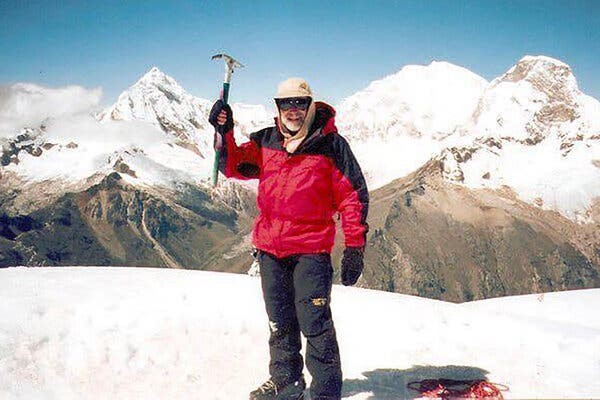 A man in a red winter coat, hat and dark sunglasses holding a climbing tool and standing on snow with several mountains behind him. 