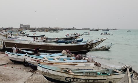 Several small artisanal fishing boats lying on a beach with a few buildings seen on a peninsula in the background